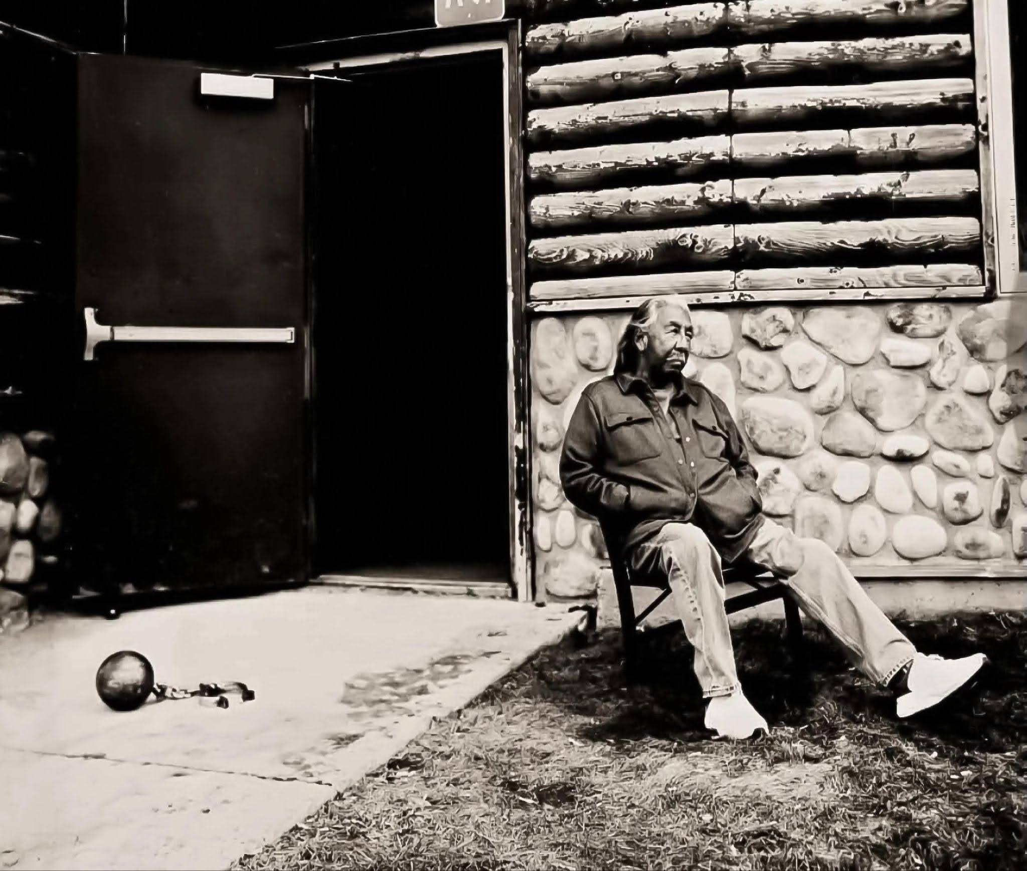 Black-and-white photograph of an older man with long hair and a beard sitting in a folding chair outside a rustic building. He wears a dark button-down shirt, light pants and white shoes, with his hands in his pockets and legs crossed. An open doorway is behind him, and a ball and chain lie on the concrete nearby.