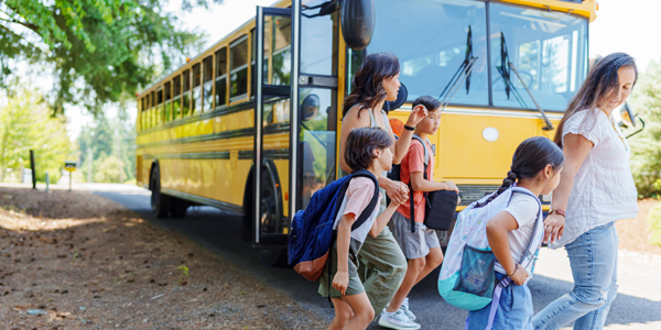 Smiling mom walk hand-in-hand with their lower-elementary aged children away from the school bus. The school bus is stopped on the side of a two-lane residential road under a tree. There are no sidewalks; the shoulders of the road are unpaved. Fly View Productions / E+ via Getty Images